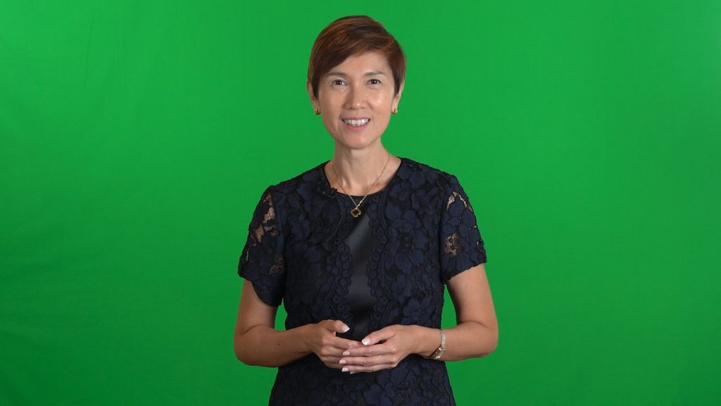 Minister Josephine Teo delivering her speech in front of a green screen backdrop.