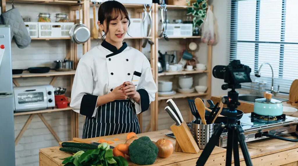 Chef in uniform filming a cooking video in a kitchen, surrounded by fresh vegetables and utensils