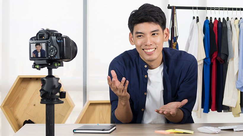 Person smiling and gesturing in front of camera at desk with notes, clothing rack, and shelf behind.
