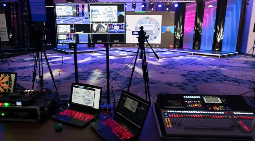 Professional event production setup with laptops, video switcher, monitors, and cameras capturing a stage presentation