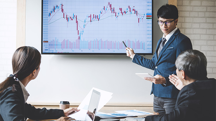 Three professionals in office meeting, presenter pointing at financial candlestick chart on large screen.