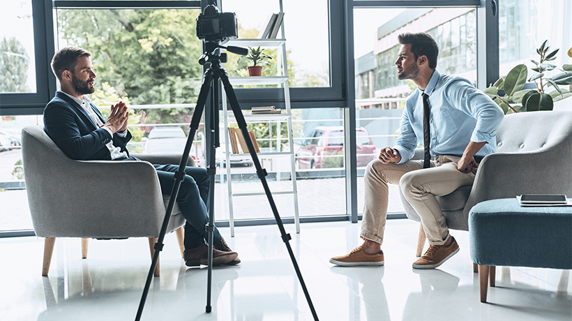 Two men having a recorded conversation in a modern studio setup with camera and natural lighting.