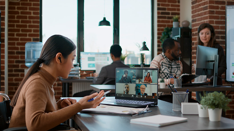 Woman video conferencing in modern office with six people on laptop screen, others working nearby.