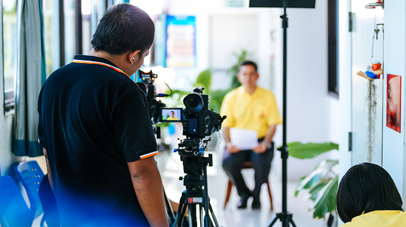 Person in yellow shirt being filmed by camera operator in indoor space with lighting equipment and decor.