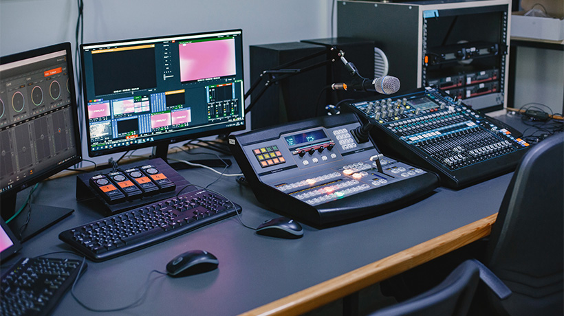 Professional studio setup with monitors, audio mixer, control panel, microphone, and editing equipment in organized workspace.