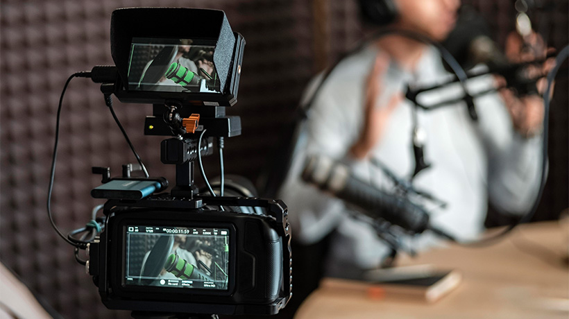 Person wearing headphones speaking into microphone while being filmed in soundproof studio with camera on tripod.