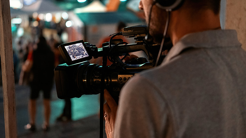 Camera operator filming outdoor event with viewfinder showing people walking near tents and booths.