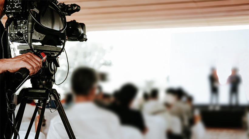 Person operating video camera on tripod, filming stage event with audience in a covered venue.
